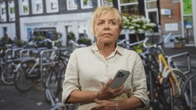 Woman holding smartphone with hands visible, wearing beige linen shirt and belt, looking up on a street by bicycle racks and a brick building; quiet contemplation. - Powered by Shutterstock - Get 15% off with code: PIKWIZARD15