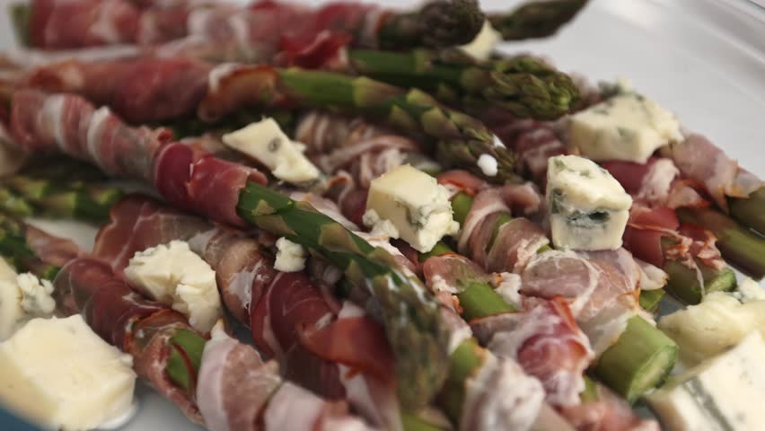 Green asparagus wrapped in bacon and arranged in a transparent oven dish, with diced gorgonzola placed between the bundles, all presented on a bright white background.
