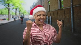 Woman points finger to camera showing hand and smile while wearing red santa hat on urban street by brick building; holiday joy. - Powered by Shutterstock - Get 15% off with code: PIKWIZARD15