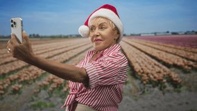 Woman holds smartphone at arm's length taking a selfie in a tulip field wearing santa hat and striped shirt; holiday joy. - Powered by Shutterstock - Get 15% off with code: PIKWIZARD15
