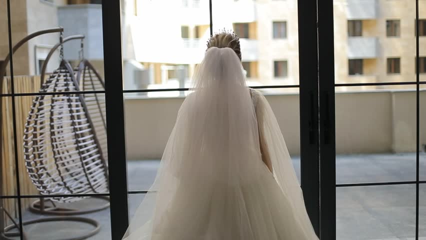 A bride, seen from behind in her beautiful white wedding dress and veil, poses with outstretched arms, looking out a large window.
