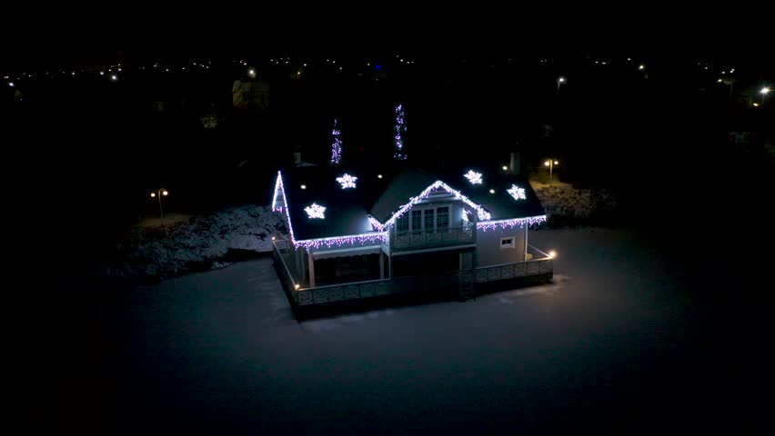 Cozy winter house decorated with bright Christmas lights and glowing snowflake ornaments captured at night during the holiday season from an elevated aerial perspective