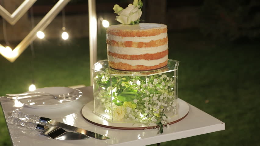 A beautiful wedding cake with white biscuit and vanilla cream, on a transparent glass base containing white flowers, set against a photo zone with lights at night.