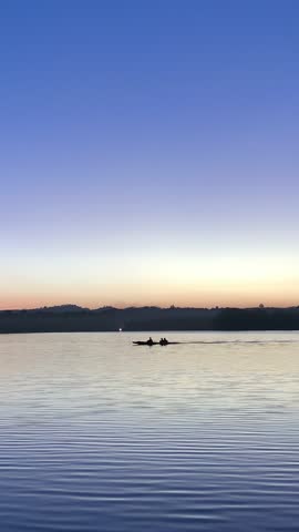 Two people rowing boat on tranquil lake at dusk with scenic landscape