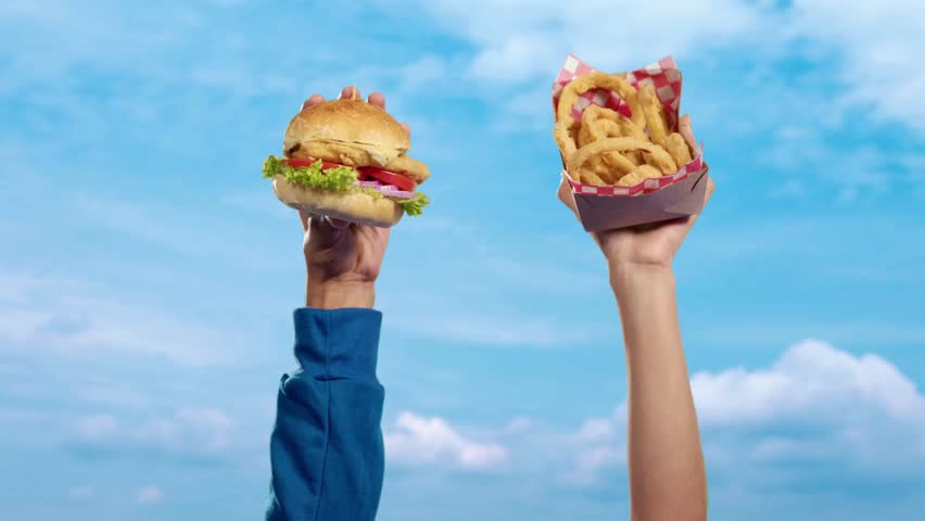 Two people holding a fresh sandwich and a box of crispy fried onion rings high into the air against a blue sky. Summer fast food advertising.