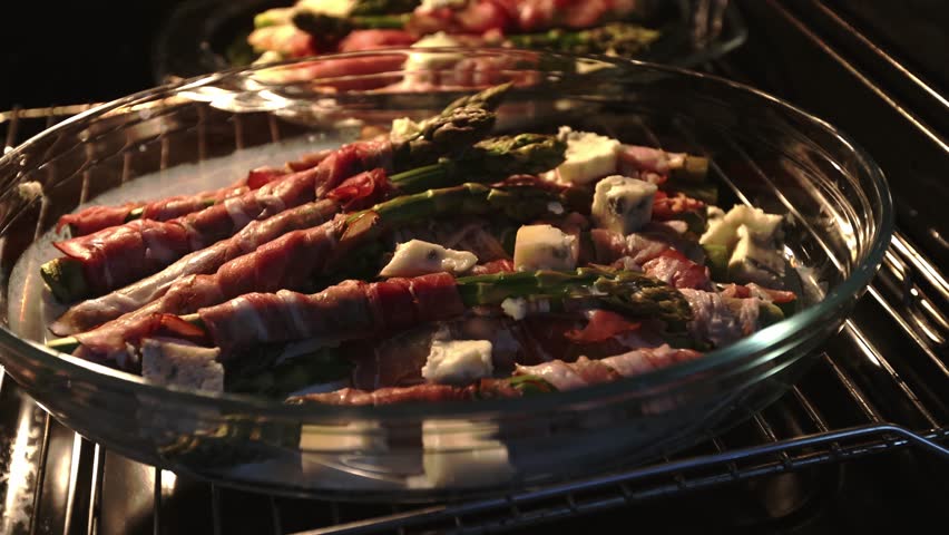 Green asparagus wrapped in bacon and arranged in a transparent oven dish, with diced gorgonzola between the bundles, shown cooking inside a heated oven with visible warm light.