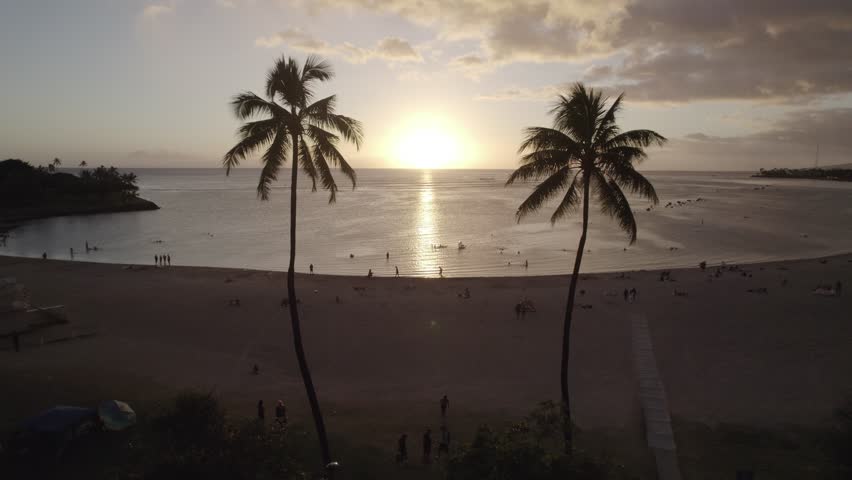 An aerial view of a tranquil Oahu beach at sunset with palm trees silhouetted against glowing Hawaiian skies