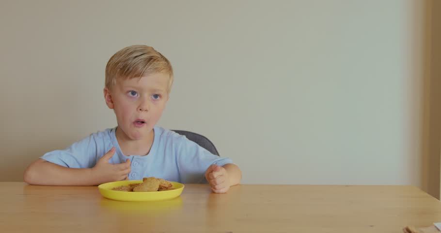A blond boy eating chicken nuggets at home, sitting at a wooden table, enjoying his snack so much that he is dancing a little.