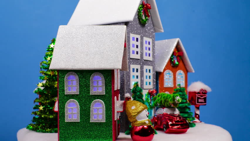 Multicolored houses with flashing lights in the windows surrounded by small Christmas trees and a snowman with a car spinning against a blue background