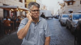 Senior man holding smartphone to ear smiling while talking on a bustling street lined with parked cars and outdoor cafes; contentment connection. - Powered by Shutterstock - Get 15% off with code: PIKWIZARD15