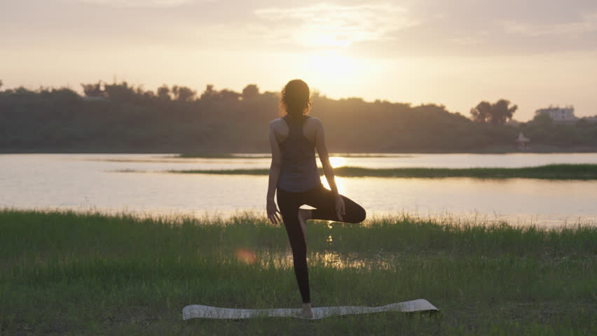 A South Asian Woman in Tree Pose (Vrksasana) at sunset while doing yoga by a lake