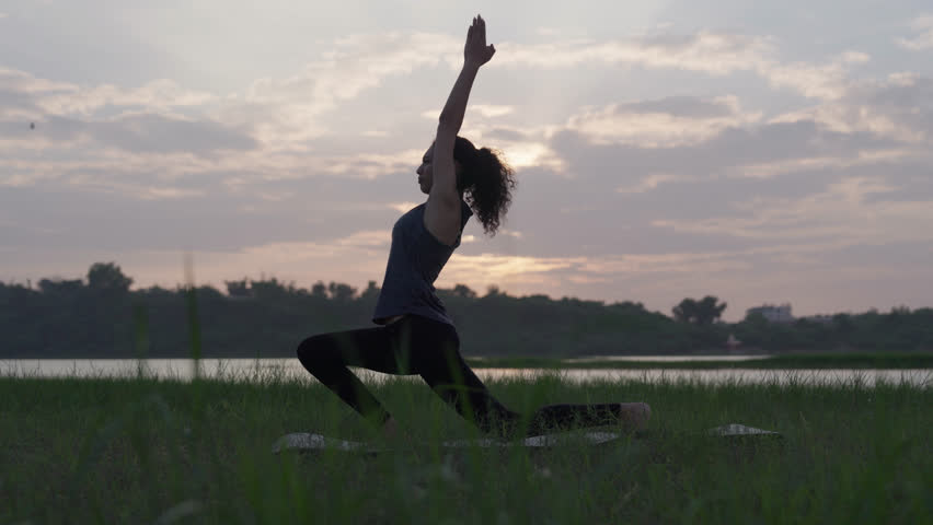 A slow-motion of a South Asian Woman in a low lunge yoga pose (Anjaneyasana) with arms raised, at sunset near a lake.