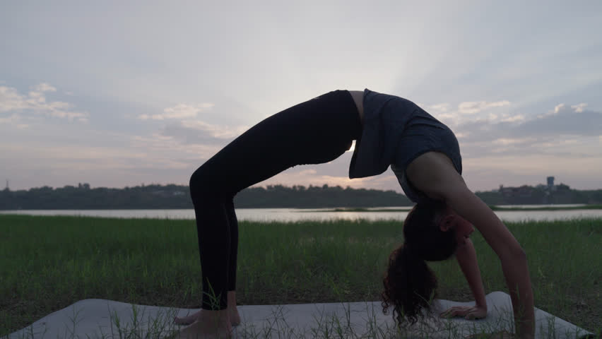 A South Asian Woman in Wheel Pose (Urdhva Dhanurasana) doing yoga on a mat outdoors by a lake at sunset.