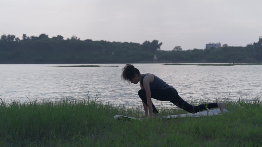 A slow-motion of a South Asian Woman in a low lunge yoga pose (Anjaneyasana) looking up, by a lake in tall grass