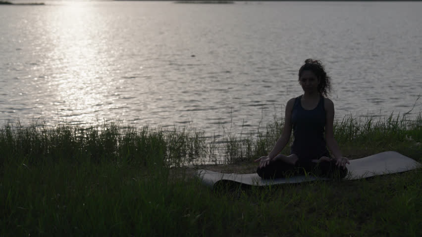 A slow-motion of South Asian Woman in Lotus Pose (Padmasana) with prayer hands silhouetted by the lake's reflection at twilight.