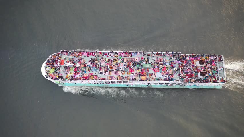 An aerial shot of crowded ferry, densely covered in people and colorful blankets, moving across dark water in Dhaka, Bangladesh