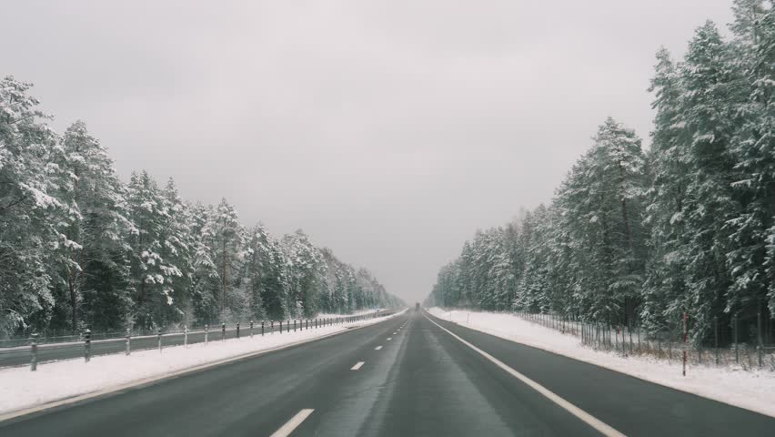 Driving on winter highway. Icy road framed by evergreen trees. Frozen asphalt path under cloudy winter sky with pines. Winter roadway bordered by snow laden pine trees beneath gray winter canopy