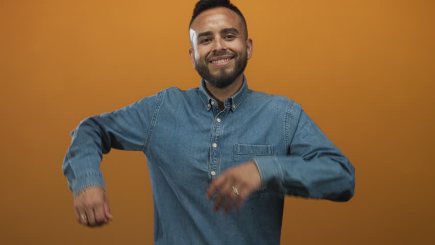 Man presenting open hands and smiling while wearing a denim shirt in orange studio; welcoming warmth friendliness.