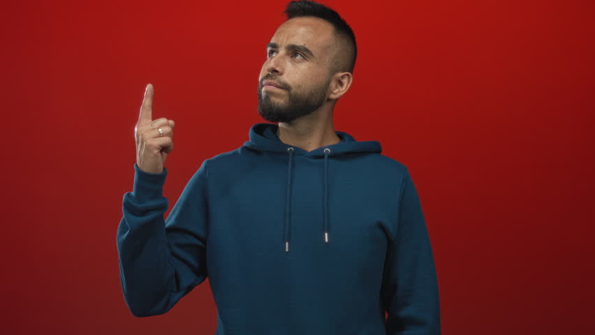 Man pointing index finger upward wearing blue hoodie in studio against red backdrop; contemplation decision resolve.