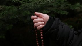 A close-up of a hand holding and running wooden rosary beads (prayer beads) against a blurred, dark green pine or fir forest background. The scene depicts spirituality, contemplation, and faith. - Powered by Shutterstock - Get 15% off with code: PIKWIZARD15
