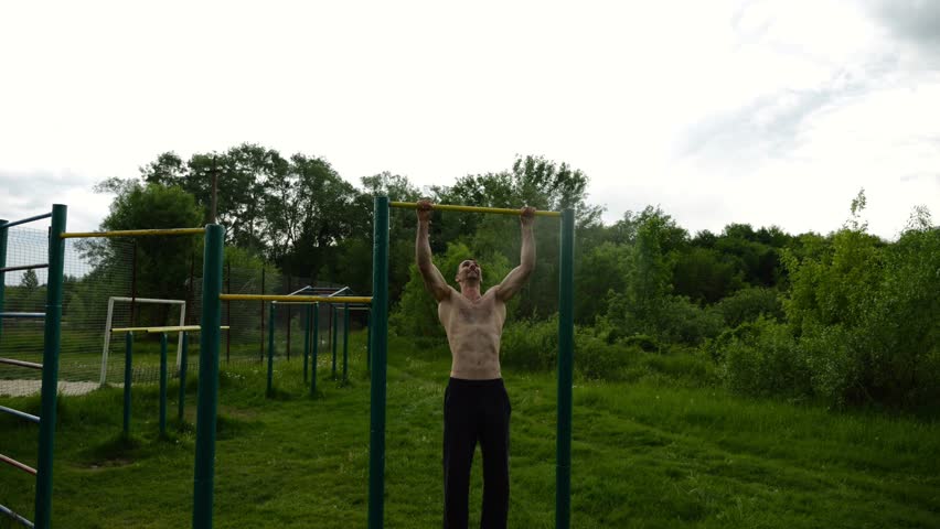 Muscular athlete doing pull ups on horizontal bar at sports ground. Young shirtless sportsman exercising at nature. Strong sporty man training outdoor. Concept of healthy and active lifestyle. Slow mo