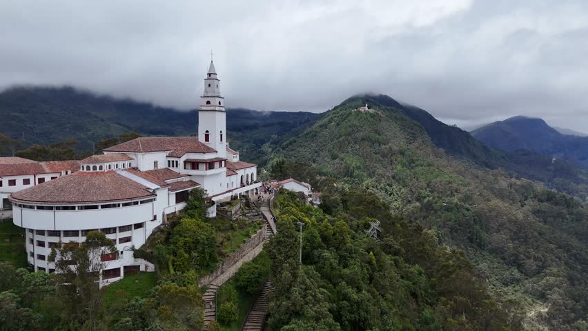 Aerial moves forward, getting closer to the white tower and main building of the historic Monserrate Sanctuary church on the mountain top, above Bogota