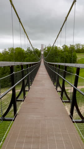 Views Across a Modern Pedestrian Suspension Bridge. Surface level views looking across a modern Suspension Bridge with views to the cloudy sky and forest and valley below.