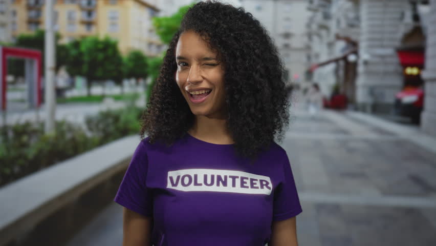 Woman in purple volunteer shirt winks while standing on urban street corner; community service happiness.