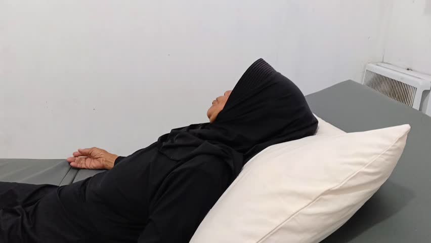 Elderly Muslim woman patient wearing a black hijab rests on a hospital bed, waking up peacefully. Concept of healthcare, recovery, faith, and quiet rest.