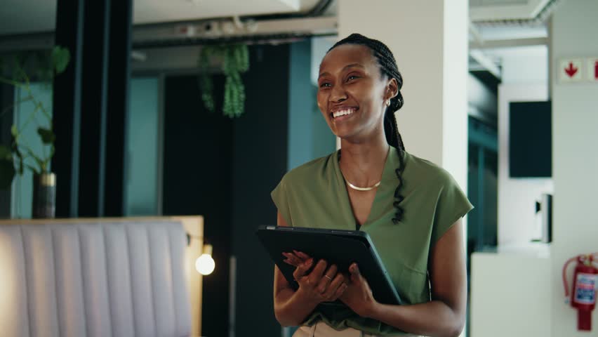 A smiling young black businesswoman with digital tablet walking in office lobby