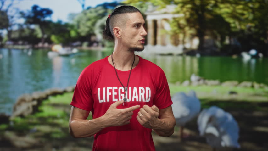 Man lifeguard in red shirt standing and gesturing with hands by green lake with trees and pavilion near building; duty vigilance.