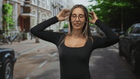 Woman with red glasses adjusting hair with hands behind head showing cleavage in black top on city street; confidence calm wellness beauty. - Powered by Shutterstock - Get 15% off with code: PIKWIZARD15