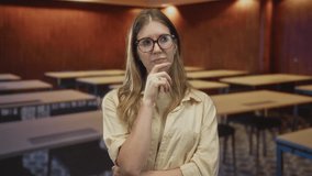 Young blonde woman wearing glasses with finger on chin for thinking in an indoor classroom; quiet contemplation. - Powered by Shutterstock - Get 15% off with code: PIKWIZARD15