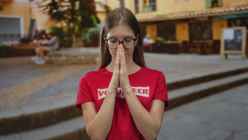 Young volunteer woman in red tshirt pressing fingertips together in front of yellow building; serenity.