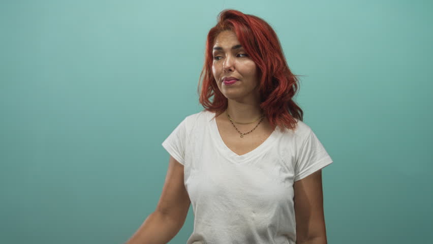 Woman with red hair wearing white t shirt pointing left with finger and making phone gesture in studio with teal backdrop; playful.