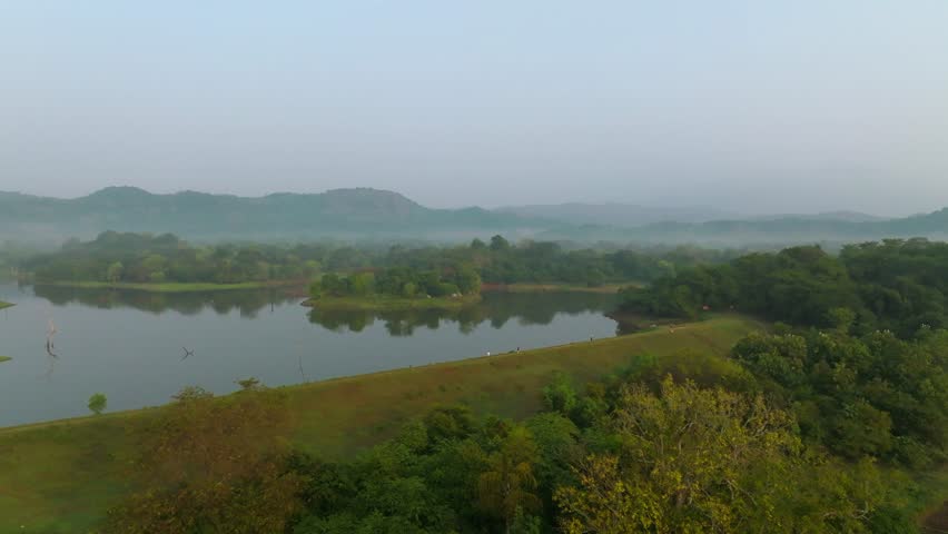Sri Lanka morning reservoir scene with a small green peninsula, calm water reflections and haze over distant hills.