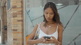 Thai woman holding smartphone with bare hands, smiling and checking phone beside building glass wall; calm confidence. - Powered by Shutterstock - Get 15% off with code: PIKWIZARD15