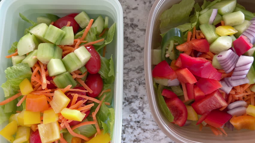A brown reusable meal prep container is filled with finely chopped salad ingredients including cucumber, red onion, red and yellow bell peppers, and shredded lettuce. All items are raw and fresh.
