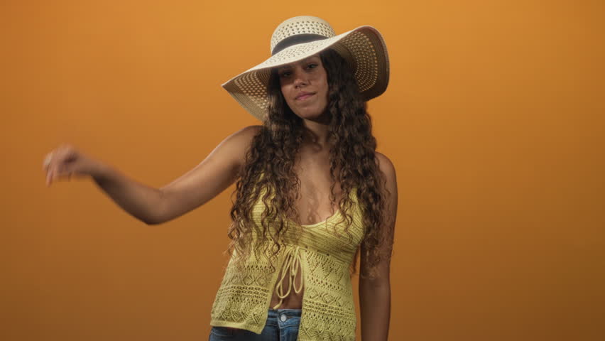 Hispanic woman sticks tongue out and raises hand to straw sunhat while wearing yellow crochet top in studio; playful.