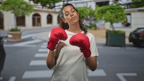 Young woman in white tshirt wearing red boxing gloves holds fists up in a guarded boxing stance on a city street crosswalk near trees and parked cars; determination strength. - Powered by Shutterstock - Get 15% off with code: PIKWIZARD15
