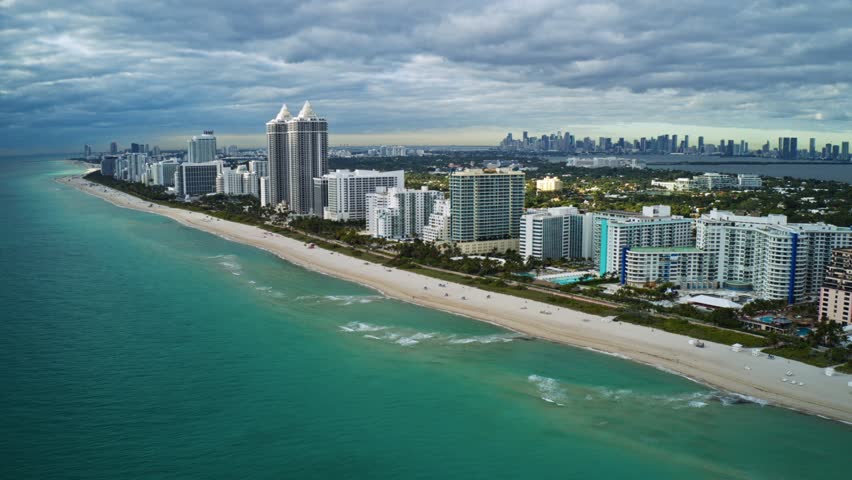 Tall oceanfront condos line the sandy shoreline of North Beach as Miami’s skyline rises in the distance, framed by turquoise Atlantic water and textured cloud cover along Florida’s southeast coast.