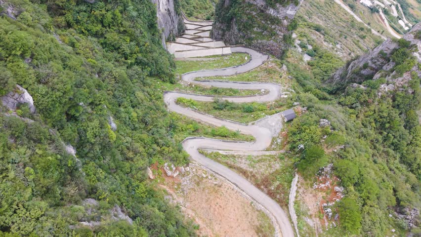 Revealing drone shot of the steep, sheer rock formations and the zigzag road carved into the cliffside at Lingpaishi, Wuxi County, China. Captures remote natural beauty and engineering.