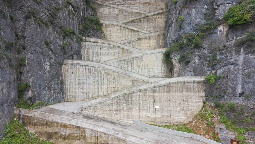 Top-down aerial view of the steep, sheer rock formations and the zigzag road carved into the cliffside at Lingpaishi, Wuxi County, China. Captures remote natural beauty and engineering. UHD.