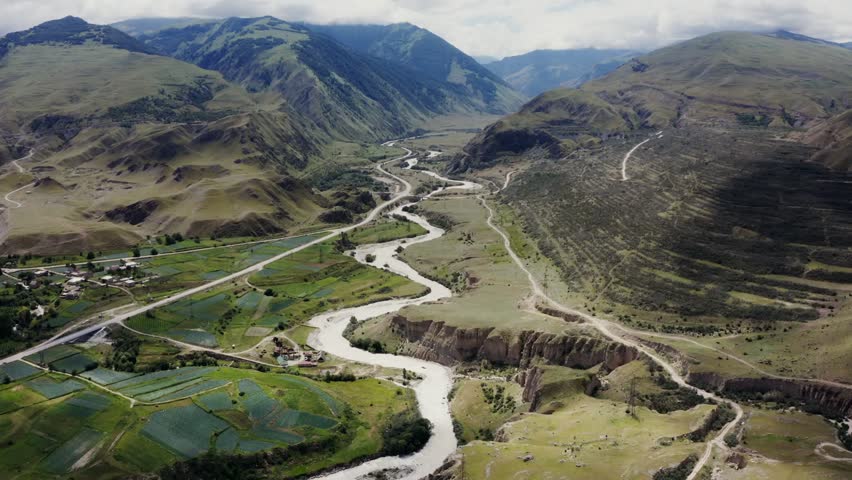 A farm with cultivated fields and a river flowing through a mountain valley, showcasing scenic slopes, agricultural patterns, and serene natural landscapes in high-altitude terrain.
