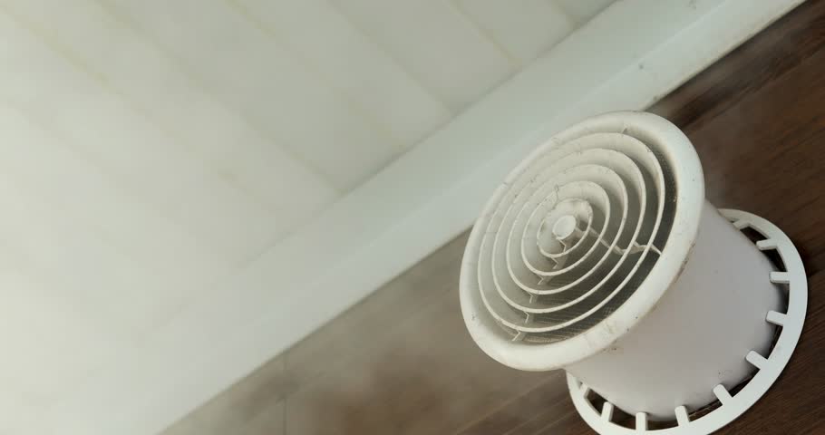 White air vent is releasing water vapor in a room. Light reflects on the wooden floor. The air vent is near the floor. This demonstrates humidity in a home.