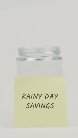 A hand deposits a coin into a glass jar featuring a yellow sticky note with Rainy Day Savings, symbolizing emergency funds, financial security, unexpected expenses, and future protection