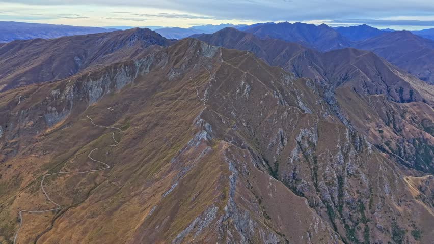 Forward drone shot following the mountain ridge of Roys Peak in Wanaka, showing the rocky slopes and winding trail under a cloudy sky.