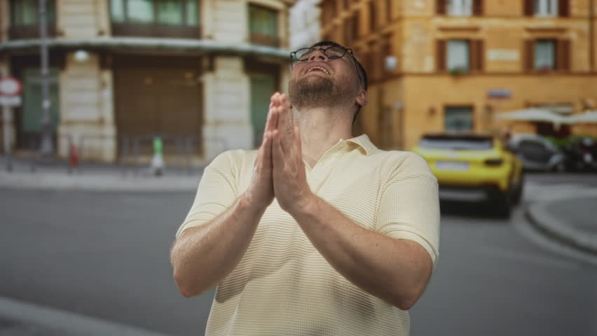 Man with hands pressed together and bowed head on a city street, wearing glasses and cream polo, palms visible; hope plea.