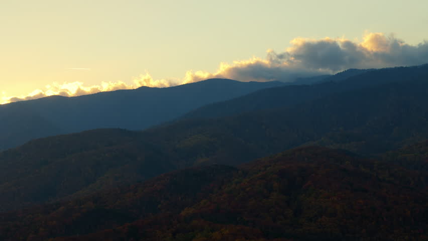 Peaceful view of Great Smoky Mountains with soft clouds rolling over the ridge. Layers of distant blue mountains blend with the golden sky