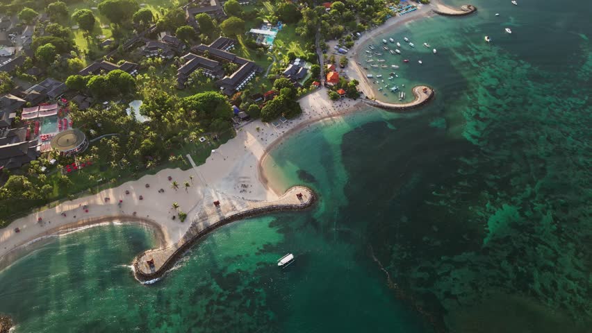 Drone footage at the Nusa Dua coastline with curved breakwaters shaping a calm reef lagoon beside a tropical beachfront where luxury resort areas line the shore creating a bright scenic ocean view